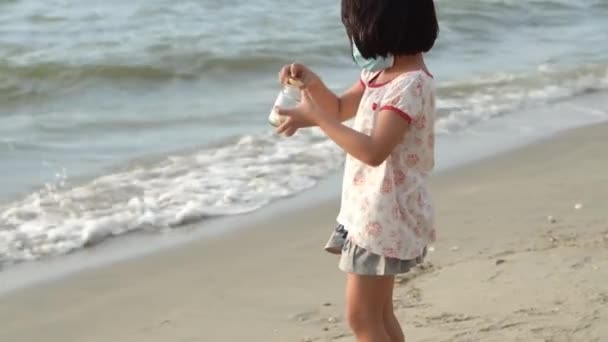 Menina com máscara facial jogar fora concha do mar quebrado na praia - Filmagem, Vídeo