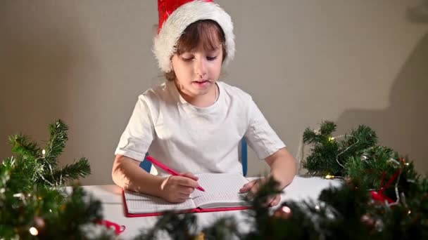 Lány gyermek fehér pólóban piros tollal és szilveszteri sapkával a fején ír egy levelet a kedves Mikulásnak az asztalnál a szülésre várva. kívánságlista ajándékok az új évre - Felvétel, videó