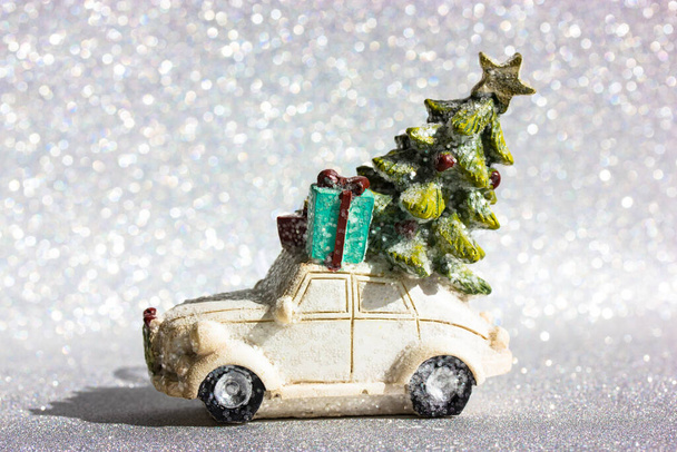 Weißes Spielzeugauto mit grünem Weihnachtsbaum auf dem Dach fährt vor silbrig glänzendem Hintergrund. Minimalistisch fröhliche Silvesternacht Hintergrund. Grußkarte, Kopierraum. Lieferung von Geschenkboxen zu Weihnachten. - Foto, Bild