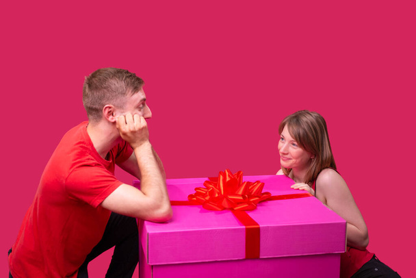 Happy young married couple man and woman in red clothes posing near the huge present box pink color looking to each other. - Fotografie, Obrázek
