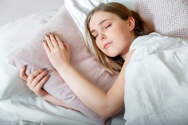 Teenager girl in pajamas sleeps in bedroom at morning time. Young Woman sleeping in bed. Portrait of blonde teen girl have healthy good sleep on white pink pillow. - Photo, image