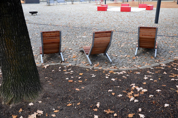 krzesło bujane na chodniku pod drzewami na placu. drewniane leżaki z drewna tropikalnego dla jednej osoby w parku. Są one komfortowo wykonane z brązowych desek, listew i stalowych ram - Zdjęcie, obraz