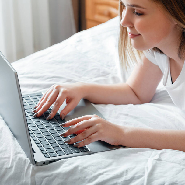Happy teen girl communicates online working remote, studying or planning her day use computer laptop in bedroom. Young woman smile typing on laptop keyboard while lying in bed at home. Square - 写真・画像