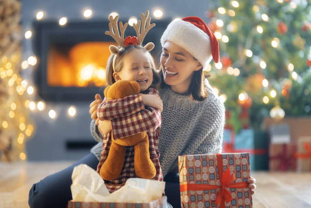 Merry Christmas and Happy Holidays. Cheerful mother and her cute daughter girl exchanging gifts. Mom and little child having fun near tree indoors. Loving family with presents in room. - Fotografie, Obrázek