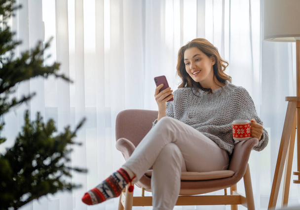 Woman is using mobile phone sitting near Christmas tree. - Фото, зображення