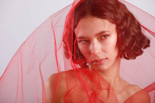 Een modieus portret van een elegante jonge vrouw met een transparante rode stof op een lichte achtergrond. - Foto, afbeelding