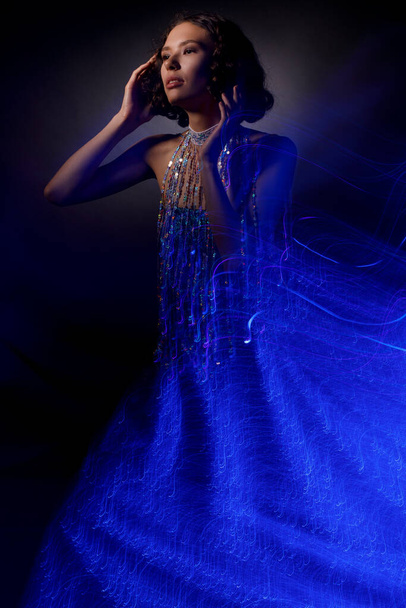 A gorgeous woman in a shiny blue dress on a dark background. - Фото, изображение