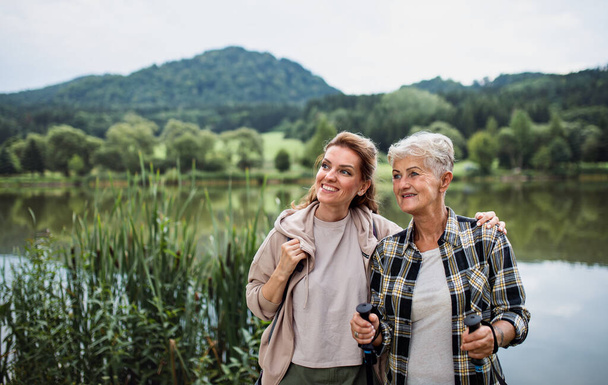 Happy senior mother embracing with adult daughter when standing by lake outdoors in nature - Photo, Image