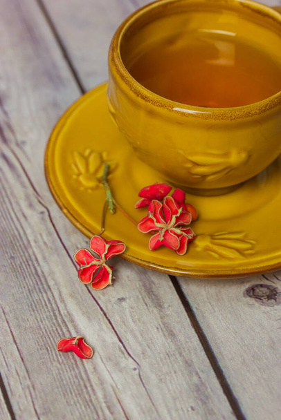 Taza de cerámica amarilla con té negro en platillo sobre mesa de madera gris. Arbusto del huso, Euonymus europaeus, Pfaffenhutchen pétalos de flores rojas. Pétalo en forma de corazón. Composición plana de naturaleza muerta. - Foto, imagen