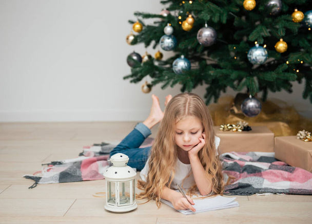 Μικρό κορίτσι γράφει γράμματα στον Άγιο Βασίλη κοντά στο χριστουγεννιάτικο δέντρο στο σαλόνι - Φωτογραφία, εικόνα