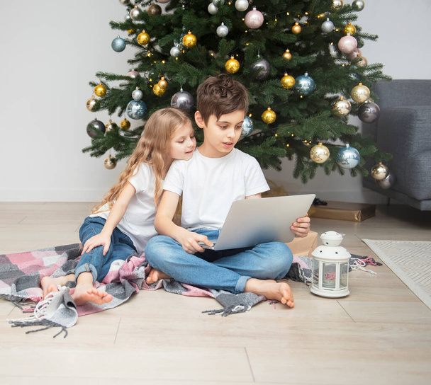 Büyük bir oturma odasında Noel ağacı. Çocuklar Noel ağacının yanında oynuyorlar. Dizüstü bilgisayarı olan bir çocuk arkadaşlarıyla iletişim kurar.. - Fotoğraf, Görsel
