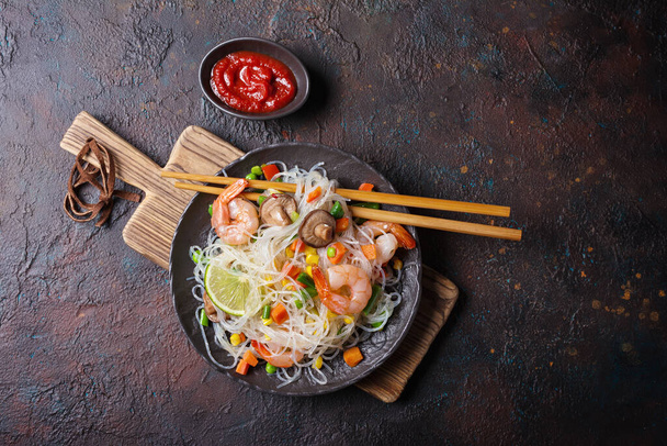 Top view of plate with japanese bean or glass noodles, tasty shrimps, shiitake mushrooms, vegetables and red sauce on dark concrete background - Photo, image