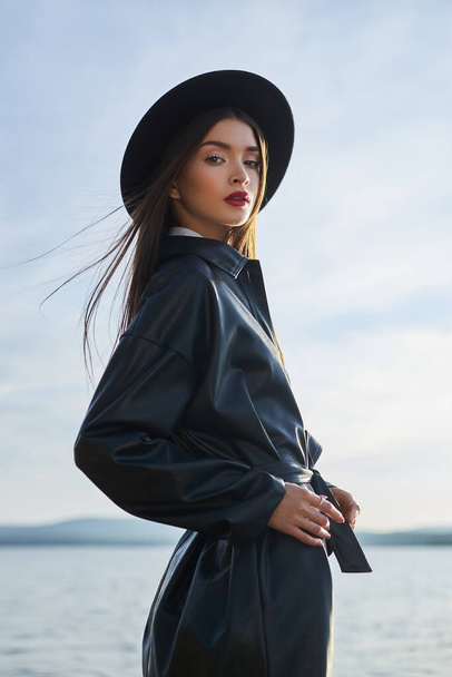 Fashion woman in black round hat and leather raincoat near lake pond. Portrait girl at sunset. Beautiful makeup and bright red lipstick on lips - Photo, image