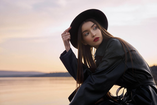 Beauty Fashion woman in black round hat and leather raincoat near lake pond. Portrait girl at sunset. Beautiful makeup and bright red lipstick on lips - Photo, image