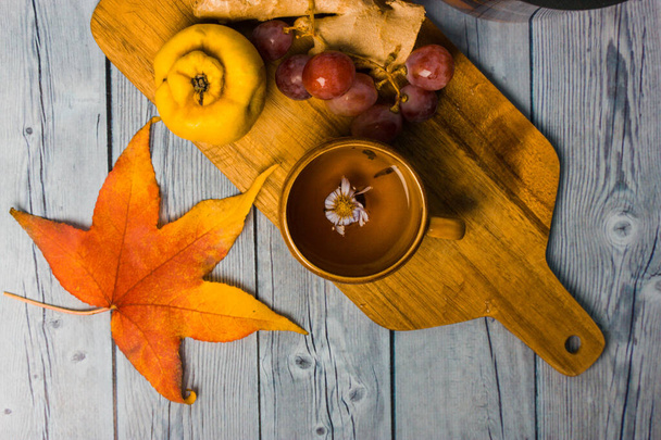 秋の静物画-ブドウの束、熟した王子、生姜の根の木製のまな板上のビューに位置しています。テーブルの上にオレンジのカエデの葉。花で装飾された熱い芳香のあるお茶を飲む黄色のカップ.  - 写真・画像