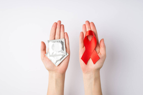 Фото от первого лица с женскими руками, держащими красную ленту и упаковки презервативов в ладонях, символ информированности о СПИДе на изолированном белом фоне - Фото, изображение