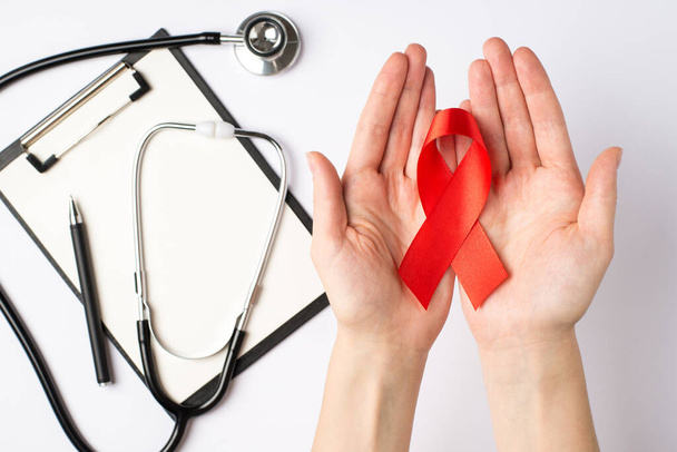 First person top view photo of female hands holding red ribbon in palms over clipboard pen and stethoscope symbol of aids awareness on isolated white background - Φωτογραφία, εικόνα