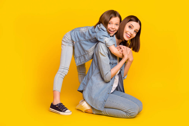 Photo of two relaxed fooling sisters wear jeans shirts smiling sitting floor embracing isolated yellow color background. - Foto, afbeelding