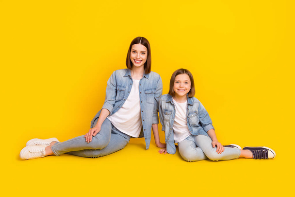 Foto de dos amigables hermanas relajadas usan camisetas vaqueras sonriendo sentado piso aislado color amarillo fondo. - Foto, Imagen