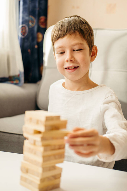 A smiling Caucasian Boy plays at home, on the table in Jenga. Board games for children and adults. A pastime without gadgets. Games during the holidays at home. Happy child plays with wooden blocks. - Fotoğraf, Görsel