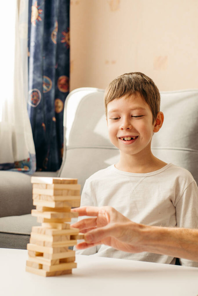 A smiling Caucasian Boy plays at home, on the table in Jenga. Board games for children and adults. A pastime without gadgets. Games during the holidays at home. Happy child plays with wooden blocks. - Фото, изображение