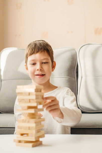 A smiling Caucasian Boy plays at home, on the table in Jenga. Board games for children and adults. A pastime without gadgets. Games during the holidays at home. Happy child plays with wooden blocks. - Photo, image