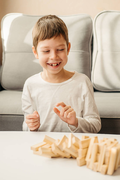 A smiling Caucasian Boy plays at home, on the table in Jenga. Board games for children and adults. A pastime without gadgets. Games during the holidays at home. Happy child plays with wooden blocks. - Foto, Imagem