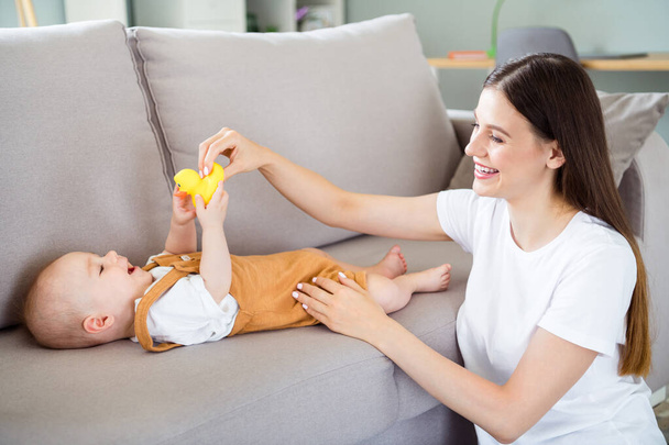 Portrait of attractive cheerful careful girl nursing baby on divan playing with toy developing at light home flat indoors. - Фото, изображение