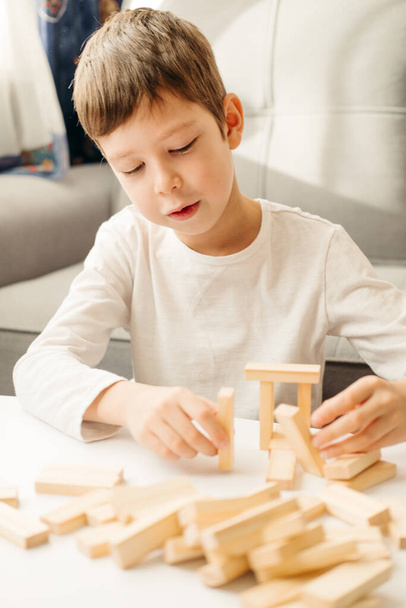 白人の少年のクローズアップは、白いTシャツで、テーブルの上に家を建てます。子供と大人のためのボードゲーム。ガジェットのない娯楽。自宅で休暇ゲーム。子供は木製のキューブで遊ぶ. - 写真・画像