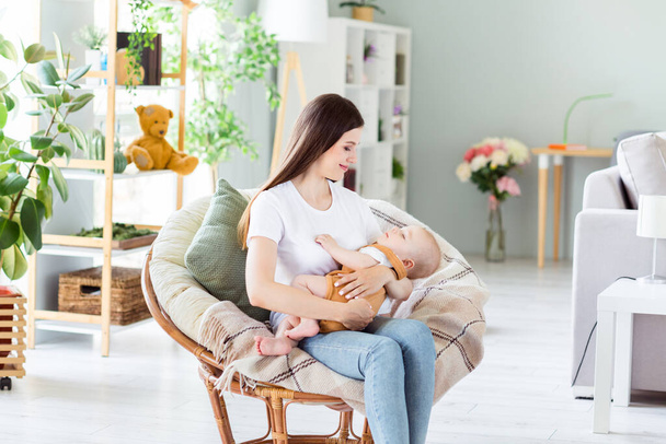 Photo of cute charming mother dressed white t-shirt sitting chair holding hands arms sleeping child inside indoors apartment room. - Foto, afbeelding