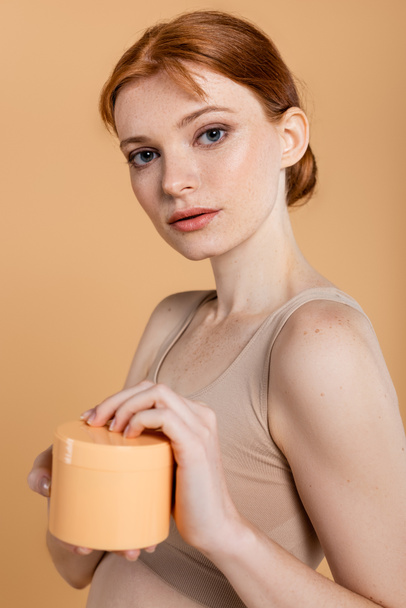 Portrait of young freckled woman holding cosmetic cream isolated on beige  - 写真・画像