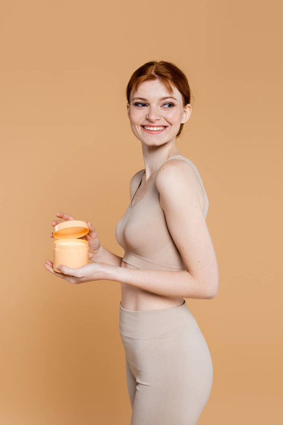 Cheerful woman with perfect skin holding container with cosmetic cream isolated on beige  - Foto, Imagem