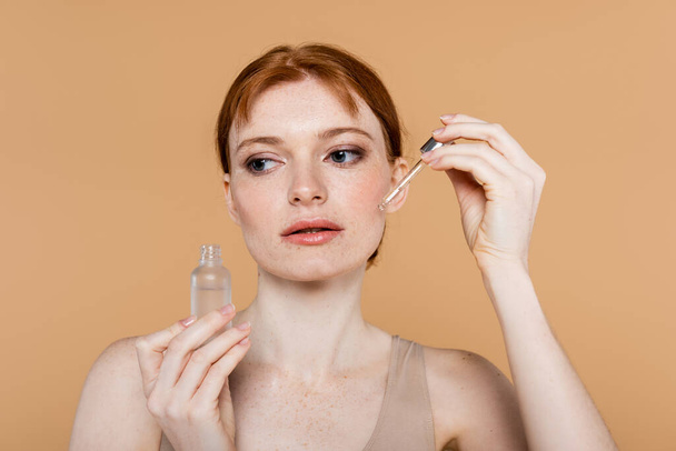 Hübsche Frau mit sommersprossiger Haut unter Anwendung kosmetischen Serums isoliert auf beige  - Foto, Bild