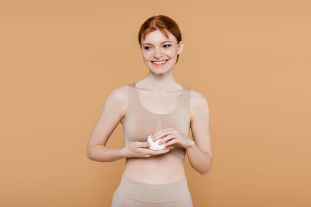Cheerful redhead woman in top holding jar with cosmetic cream isolated on beige  - Foto, immagini