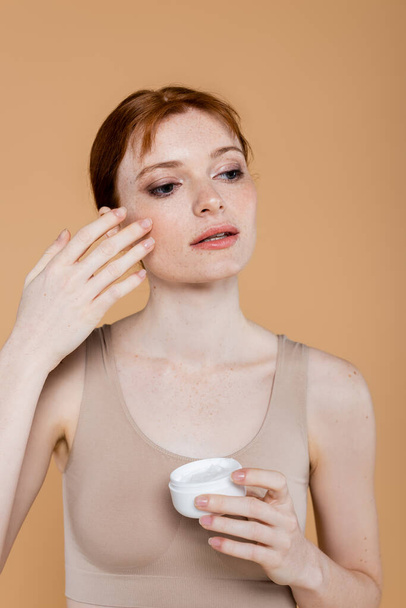 Young woman in top applying cream on face isolated on beige  - Fotó, kép