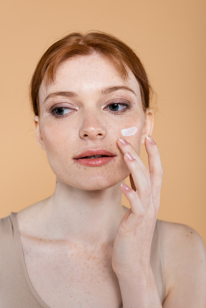 Portrait of young woman applying cosmetic cream on face isolated on beige  - Fotó, kép