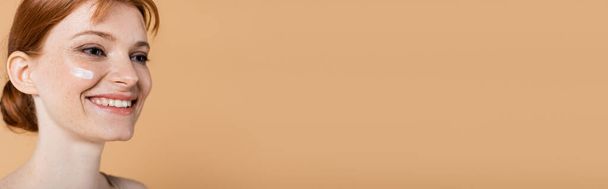 Souriante femme taches de rousseur avec crème cosmétique sur le visage regardant loin isolé sur beige avec espace de copie, bannière  - Photo, image