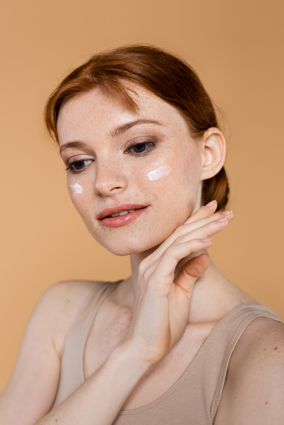 Pretty red haired woman with cosmetic cream on face posing isolated on beige  - Фото, изображение