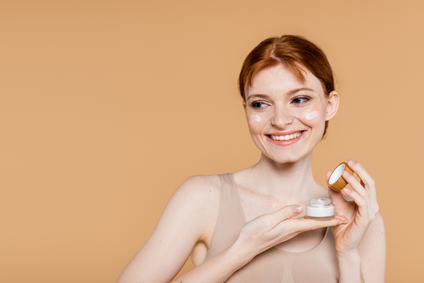 Mujer feliz sosteniendo contenedor con crema cosmética y mirando hacia otro lado aislado en beige  - Foto, imagen