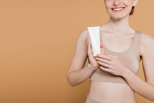 Oříznutý pohled na usmívající se žena v horní drží trubice s kosmetickou krém izolované na béžové  - Fotografie, Obrázek