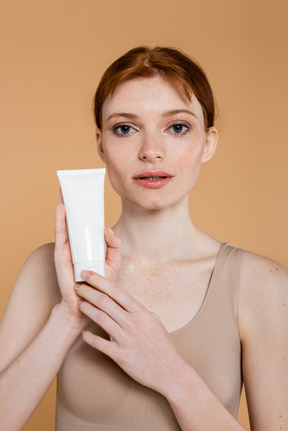 Pretty red haired woman holding tube with cosmetic cream and looking at camera isolated on beige  - Foto, immagini