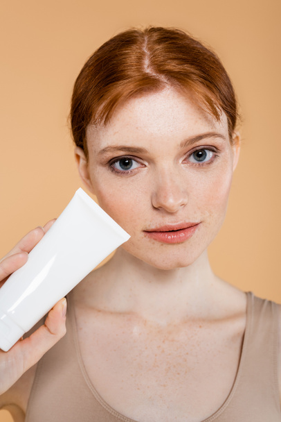 Portrait of freckled woman holding tube with cream isolated on beige  - Photo, image