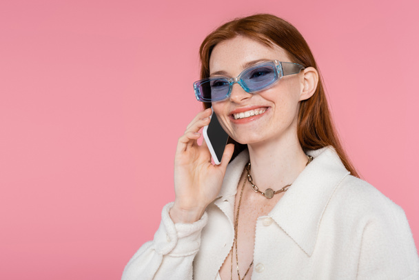 Stylish red haired woman in sunglasses talking on smartphone isolated on pink - Foto, afbeelding