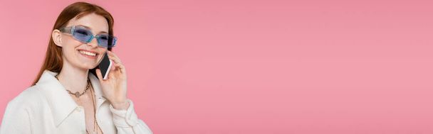 Pretty red haired woman in sunglasses talking on cellphone isolated on pink, banner  - Fotó, kép
