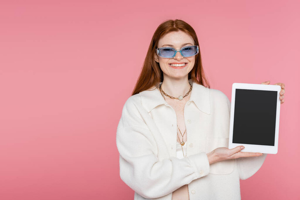 Smiling red haired woman in sunglasses holding digital tablet with blank screen isolated on pink - 写真・画像