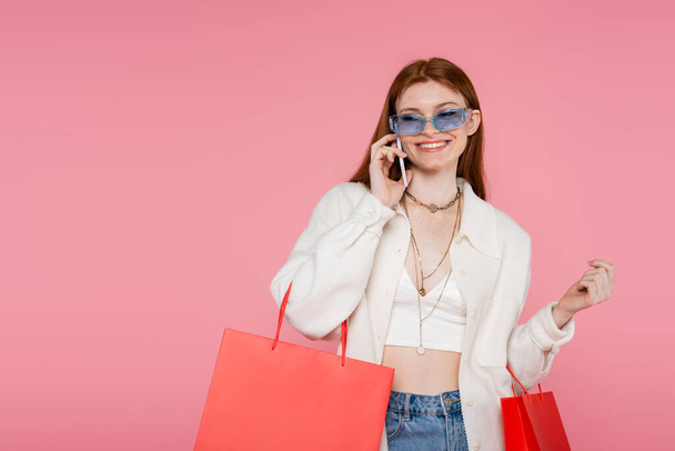 Stylish woman in sunglasses holding shopping bags and talking on smartphone isolated on pink - Photo, Image