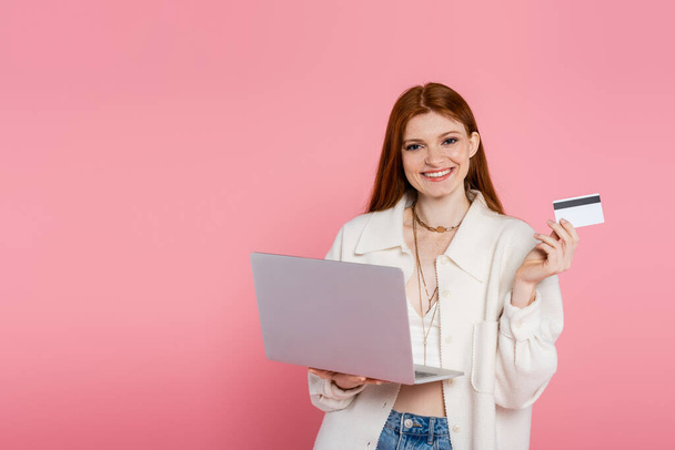 Stylish red haired woman holding credit card and laptop isolated on pink - Фото, зображення