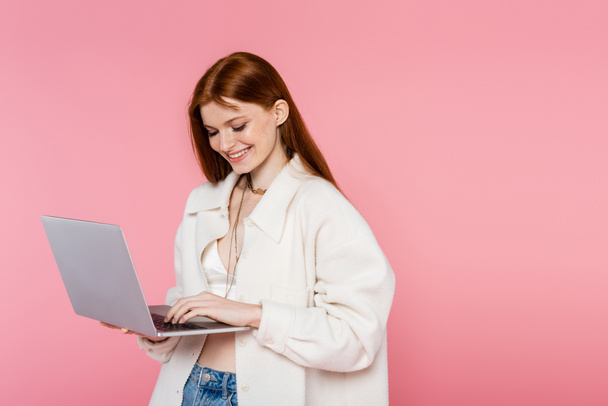 Smiling red haired woman in jacket using laptop isolated on pink - Valokuva, kuva