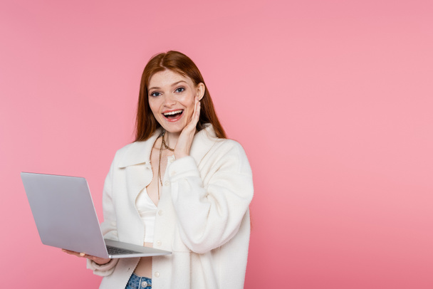 Aufgeregt rothaarige Frau in Jacke mit Laptop und Blick in die Kamera isoliert auf rosa  - Foto, Bild