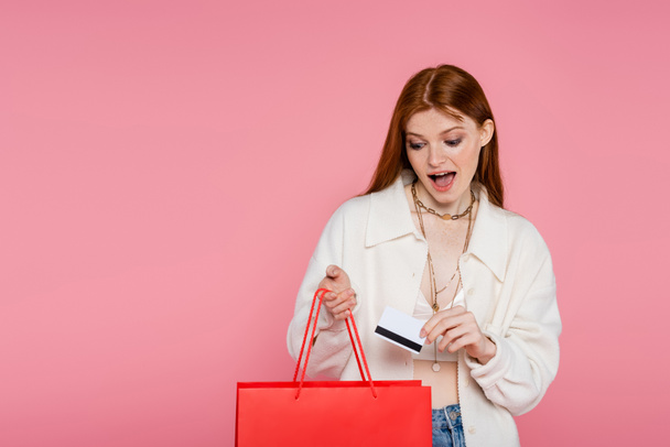 Εκπληκτική κοκκινομάλλα γυναίκα που κρατάει πιστωτική κάρτα και κοιτάζει την τσάντα με τα ψώνια απομονωμένη σε ροζ χρώμα.  - Φωτογραφία, εικόνα